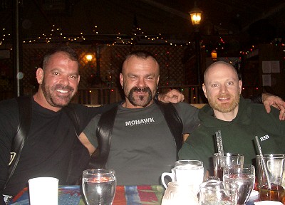 Michael, Bo and John sitting on one side of a restaurant dinner table with their arms around each other