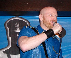 John on stage wearing a blue leather shirt, talking into a microphone
