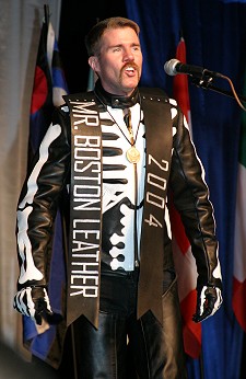 Scott in full leather, wearing his Mr Boston Leather sash, speaking into a microphone onstage at IML