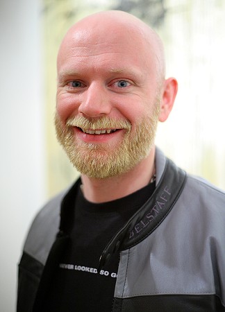 John wearing a black t-shirt and grey leather jacket, smiling at the camera