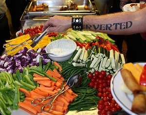 An outstretched arm at the buffet table, with PERVERTED tattooed up the forearm