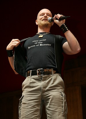 John on stage, pulling his waistcoat open to reveal a t-shirt which says slave cerise of the house of hampster