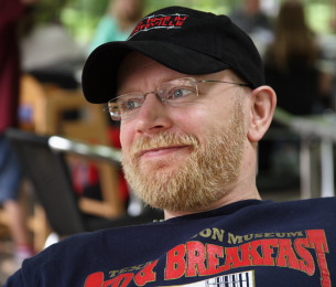 Closeup of John's smiling face, he's wearing a baseball cap and glasses