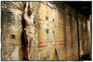 Heavily tattoed model wearing a loincloth hanging by his hands from a short pipe sticking out of a wall