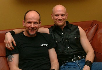 John with his arm around Kurt, both sitting on a brown leather sofa