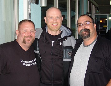 Mid-shot of three men outside, stood close together and smiling at the camera