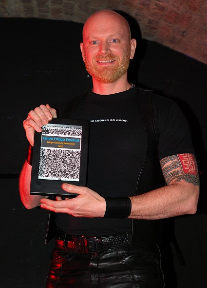John wearing black leather jeans and waistcoat and a black cotton t-shirt, holding up the award he won