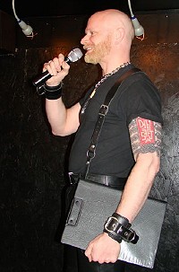 John standing sideways on stage, holding a clipboard and microphone